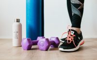 Crop sportswoman wearing sneakers and black leggings standing with sport mat on wooden floor near dumbbells and water bottle before exercising indoors